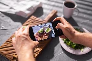 as mãos tiram fotos no smartphone de dois lindos sanduíches de creme azedo e abacate a bordo sobre a mesa. mídia social e conceito de comida