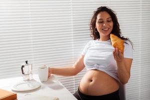 alegre grávida linda jovem comendo croissant e bebendo chá durante o café da manhã. conceito de manhã agradável e expectativa de conhecer o bebê. copyspace foto