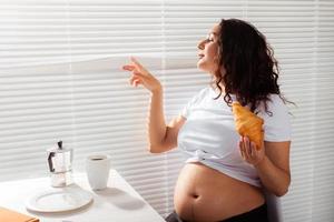 mulher bonita jovem grávida feliz comendo croissant e olhando através das persianas durante o café da manhã. conceito de manhã agradável e atitude positiva durante a gravidez foto