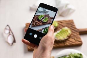 as mãos tiram fotos no smartphone de dois lindos sanduíches de creme azedo e abacate a bordo sobre a mesa. mídia social e conceito de comida