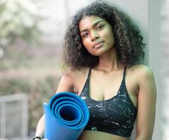 mulher de cabelo afro feliz em pé ao ar livre se preparando para treinamento de ioga, exercício ou treino. retrato de jovem mulher atlética em roupas esportivas segurando esteira para relaxamento e exercício físico. copie o espaço foto