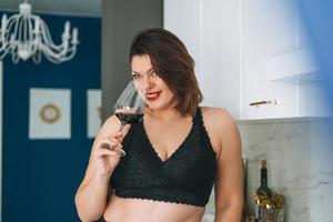 linda feliz morena sensual jovem mulher plus size positiva com um copo de vinho tinto curtindo sua vida em casa foto