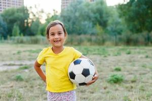 foto ao ar livre de menina bonitinha, apoiando-se na bola de futebol na grama verde