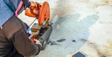 o trabalhador na roupa de trabalho usa um cortador de aço elétrico. cortando grandes barras de aço no canteiro de obras foto