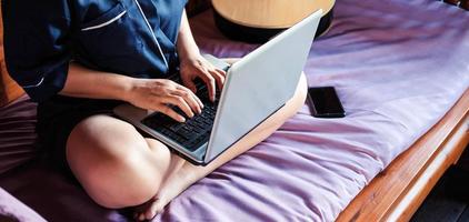 jovem mulher asiática moderna em uma roupa casual e óculos, sentada em uma mesa com um notebook e trabalhando em um trabalho em casa foto