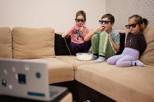 três crianças sentadas na sala de estar, usam óculos 3d assistindo filme ou desenho animado. foto