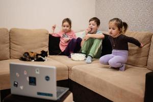 três crianças com gatinho sentado na sala assistindo filme ou desenho animado do laptop. foto