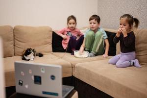 três crianças com gatinho sentado na sala assistindo filme ou desenho animado do laptop. foto