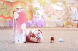 várias latas de spray usadas com tinta rosa e branca e tampas para pulverizar tinta sob pressão estão no asfalto perto da parede pintada em desenhos de graffiti coloridos foto