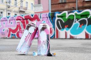 várias latas de spray usadas com tinta rosa e branca e tampas para pulverizar tinta sob pressão estão no asfalto perto da parede pintada em desenhos de graffiti coloridos foto