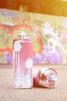 várias latas de spray usadas com tinta rosa e branca e tampas para pulverizar tinta sob pressão estão no asfalto perto da parede pintada em desenhos de graffiti coloridos foto