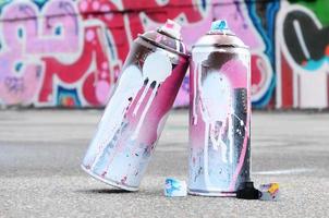 várias latas de spray usadas com tinta rosa e branca e tampas para pulverizar tinta sob pressão estão no asfalto perto da parede pintada em desenhos de graffiti coloridos foto