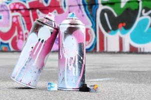 várias latas de spray usadas com tinta rosa e branca e tampas para pulverizar tinta sob pressão estão no asfalto perto da parede pintada em desenhos de graffiti coloridos foto