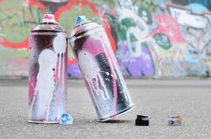 várias latas de spray usadas com tinta rosa e branca e tampas para pulverizar tinta sob pressão estão no asfalto perto da parede pintada em desenhos de graffiti coloridos foto