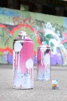 várias latas de spray usadas com tinta rosa e branca e tampas para pulverizar tinta sob pressão estão no asfalto perto da parede pintada em desenhos de graffiti coloridos foto