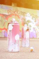 várias latas de spray usadas com tinta rosa e branca e tampas para pulverizar tinta sob pressão estão no asfalto perto da parede pintada em desenhos de graffiti coloridos foto