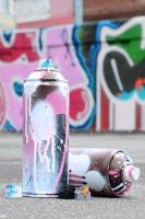 várias latas de spray usadas com tinta rosa e branca e tampas para pulverizar tinta sob pressão estão no asfalto perto da parede pintada em desenhos de graffiti coloridos foto