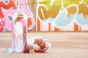 várias latas de spray usadas com tinta rosa e branca e tampas para pulverizar tinta sob pressão estão no asfalto perto da parede pintada em desenhos de graffiti coloridos foto