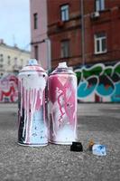 várias latas de spray usadas com tinta rosa e branca e tampas para pulverizar tinta sob pressão estão no asfalto perto da parede pintada em desenhos de graffiti coloridos foto