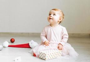 menina bonitinha de vestido rosa com saia tutu e chapéu de Papai Noel com caixa de presente presente sentada no chão na sala com brinquedos de natal em casa, feliz ano novo foto