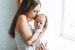 mãe jovem com cabelo comprido, segurando o bebê nas mãos perto da janela em casa foto