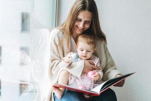 menina bonitinha de vestido rosa com sua mãe jovem lendo livro no quarto em casa foto