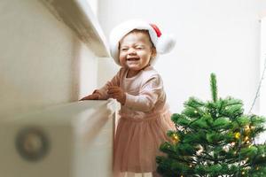 menina bonitinha de vestido rosa e chapéu de papai noel no quarto com árvore de natal em casa, feliz ano novo foto