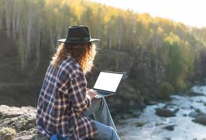 jovem mulher bonita com cabelos cacheados no chapéu de feltro e camisa xadrez usando laptop no fundo de vista mágica de montanhas e rio, caminhadas na natureza outono, conceito de freelance foto
