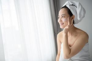 feliz jovem asiática aplicando loções faciais enquanto usava uma toalha e tocando seu rosto. maquiagem diária e cuidados com a pele foto