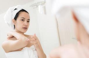 feliz jovem mulher asiática aplicando loções em seu corpo no banheiro foto