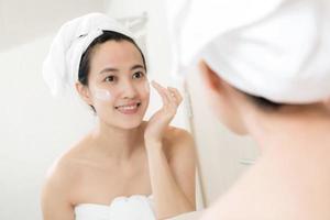 feliz jovem asiática aplicando loções faciais enquanto usava uma toalha e tocando seu rosto no banheiro foto