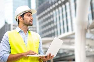 homem caucasiano usa laptop para trabalhar e usar um colete amarelo e capacete no canteiro de obras da cidade. foto