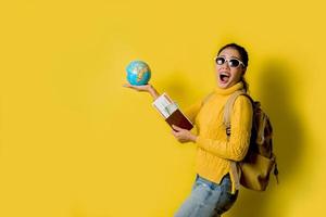 viajante de mulher com mala, segurando o globo de bola na mão com passaporte e passagem em fundo amarelo. retrato de menina feliz sorridente com espaço copie o texto. mochila de viagem foto