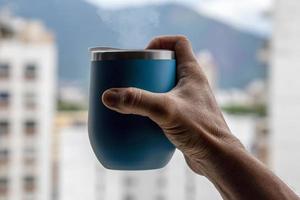 mão segurando uma caneca de café saindo da fumaça com fundo de ambiente urbano foto