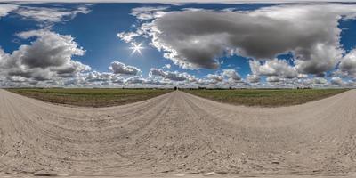 panorama full hdri 360 em estrada de cascalho sem trânsito entre campos com céu nublado e nuvens fofas brancas em projeção esférica sem costura equiretangular, pode ser usado como substituto do céu em panoramas foto