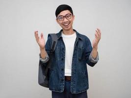 homem asiático positivo jeans camisa usar óculos bolsa de transporte mostrar as mãos para cima sorriso alegre isolado foto