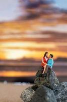 pessoas em miniatura, casal sentado em uma praia do mar com fundo pôr do sol foto
