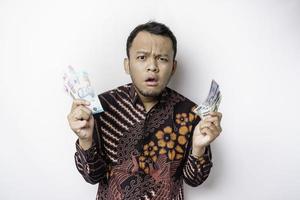 um jovem asiático confuso está vestindo camisa batik e segurando dinheiro em rupias indonésias isolado por fundo branco foto