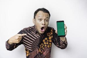 homem asiático chocado vestindo camisa batik e mostrando tela verde em seu telefone, isolado por fundo branco foto