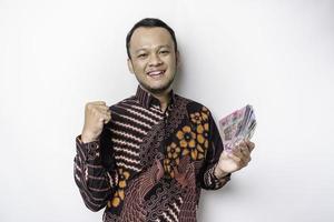 um jovem asiático feliz está vestindo camisa batik e segurando dinheiro em rupias indonésias isolado por fundo branco foto