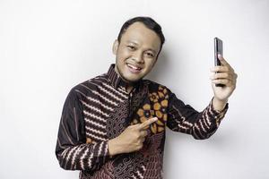 um retrato de um homem asiático feliz vestindo camisa batik e segurando seu telefone, isolado por fundo branco foto