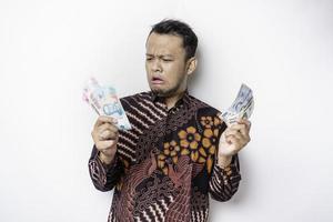 um jovem asiático confuso está vestindo camisa batik e segurando dinheiro em rupias indonésias isolado por fundo branco foto