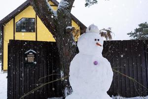 boneco de neve engraçado em um chapéu e cachecol no fundo de uma casa amarela no quintal. inverno, entretenimento de inverno, queda de neve foto