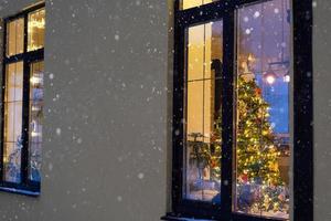 janela festiva aconchegante da casa do lado de fora com a luz quente das guirlandas de luzes de fada dentro - comemore o natal e o ano novo em um lar aconchegante. árvore de natal, bokeh, neve em pinheiros e queda de neve foto