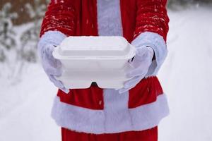 recipientes de serviço de entrega de comida nas mãos do papai noel ao ar livre na neve. promoção de véspera de natal. pedido quente pronto, caixa plástica descartável. catering de férias de ano novo. copie o espaço, simular foto