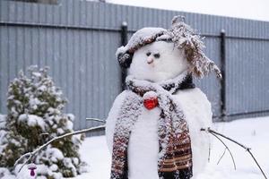 boneco de neve engraçado em um chapéu e cachecol no fundo de uma casa amarela no quintal. inverno, entretenimento de inverno, queda de neve foto