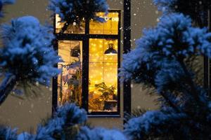 janela festiva aconchegante da casa do lado de fora com a luz quente das guirlandas de luzes de fada dentro - comemore o natal e o ano novo em um lar aconchegante. árvore de natal, bokeh, neve em pinheiros e queda de neve foto