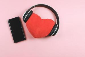 postura plana de travesseiro de coração vermelho coberto com fones de ouvido e celular de tela preta em branco no fundo rosa. canções de amor ou conceito de podcast. foto
