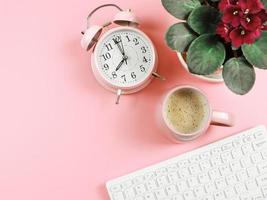 configuração plana de teclado de computador, xícara de café rosa, despertador vintage rosa e violetas africanas em vaso de planta em fundo rosa com espaço de cópia. foto