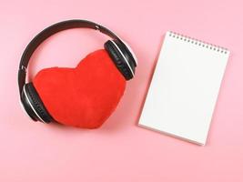 postura plana de travesseiro de coração vermelho coberto com fones de ouvido, página em branco aberta caderno em fundo rosa. canções de amor ou conceito de podcast. foto